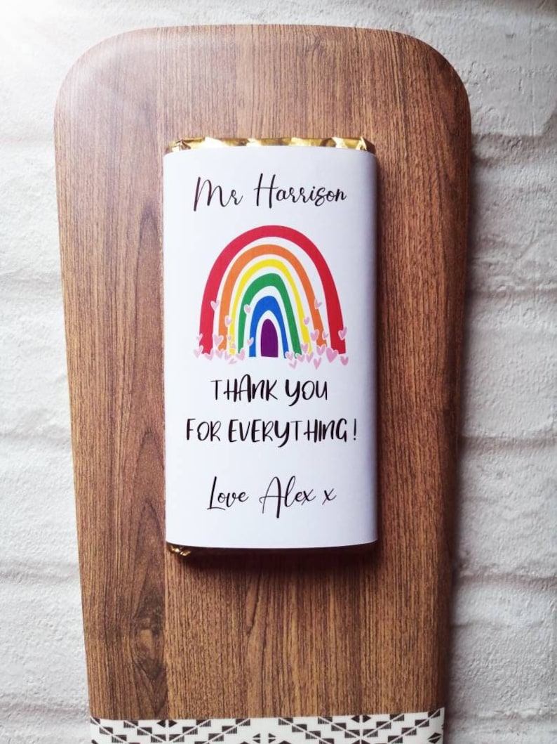 Op de afbeelding: Een chocoladereep met een witte wikkel met een regenboogontwerp en de tekst "Mr Harrison, Thank You For Everything! Love Alex x." De reep ligt op een houten snijplank met een decoratief lint.