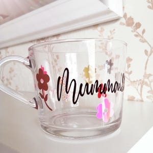 May include: A clear glass mug with a black handle and the word "Mumma" written in black script. The mug is decorated with colorful flowers.