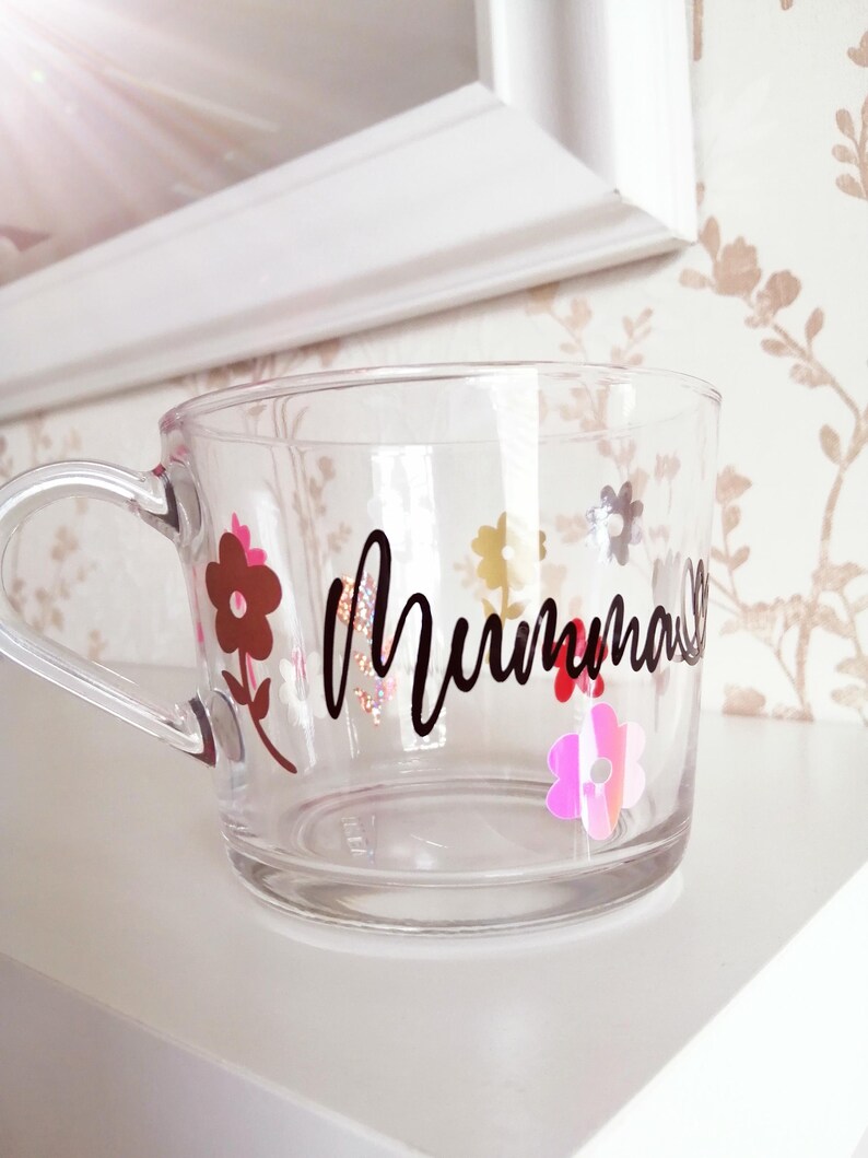 May include: A clear glass mug with a black handle and the word "Mumma" written in black script. The mug is decorated with colorful flowers.