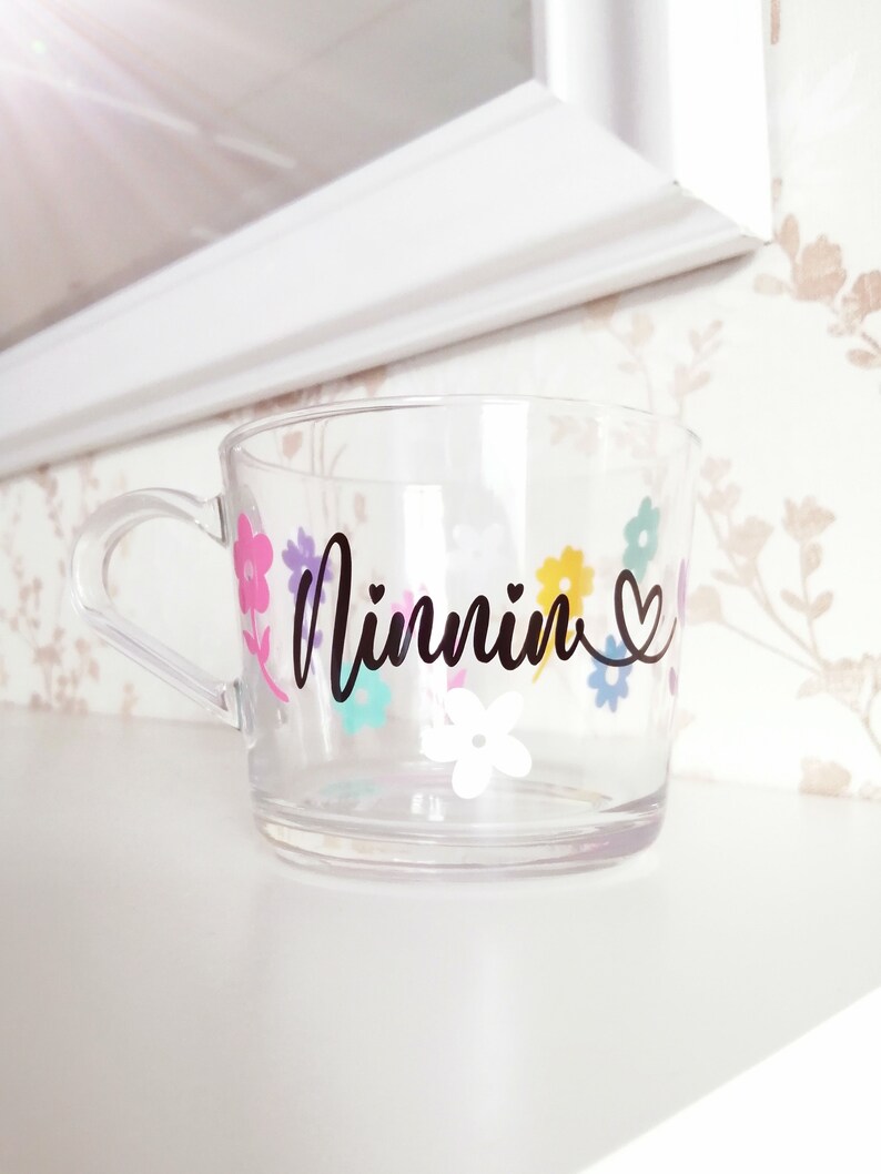 May include: Clear plastic cup with a floral design and the word "Minnin" written in black cursive. The cup has a handle and a heart symbol next to the name.