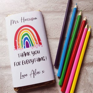 Op de afbeelding: Een chocoladereep verpakt in wit papier met een regenboogontwerp en de tekst "Thank You For Everything!" en "Love Alex x". Daarnaast liggen zes kleurpotloden in paars, blauw, groen, rood, roze en geel.