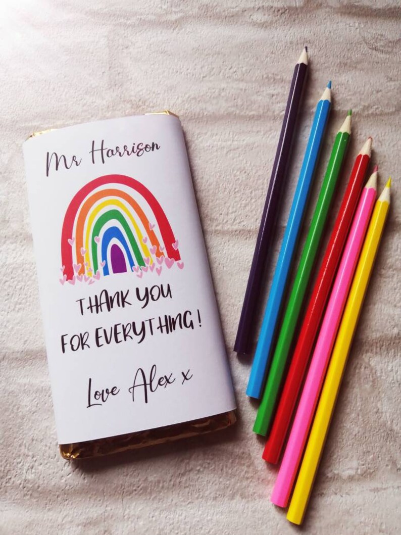 Op de afbeelding: Een chocoladereep verpakt in wit papier met een regenboogontwerp en de tekst "Thank You For Everything!" en "Love Alex x". Daarnaast liggen zes kleurpotloden in paars, blauw, groen, rood, roze en geel.