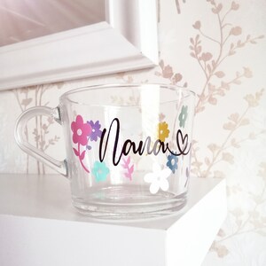 May include: A clear glass mug with a black floral design and the word "Mama" in black script. The flowers are pink, purple, yellow, blue, and white.