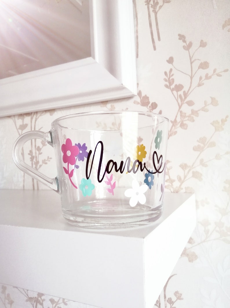 May include: A clear glass mug with a black floral design and the word "Mama" in black script. The flowers are pink, purple, yellow, blue, and white.