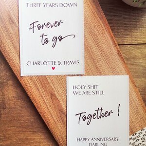 Pu&ograve; includere: Due carte bianche con testo nero su una superficie di legno. La carta superiore dice "Tre anni gi&ugrave;, Per sempre andare, Charlotte & Travis". La carta inferiore dice "Porca miseria, siamo ancora insieme! Buon anniversario, tesoro".