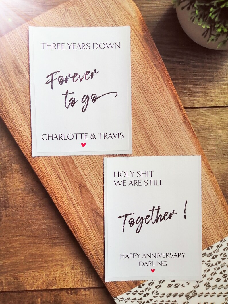 Pu&ograve; includere: Due carte bianche con testo nero su una superficie di legno. La carta superiore dice "Tre anni gi&ugrave;, Per sempre andare, Charlotte & Travis". La carta inferiore dice "Porca miseria, siamo ancora insieme! Buon anniversario, tesoro".