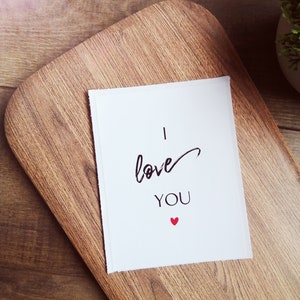 Pu&ograve; includere: Un biglietto bianco con testo nero che dice "Ti amo" e un piccolo cuore rosso.