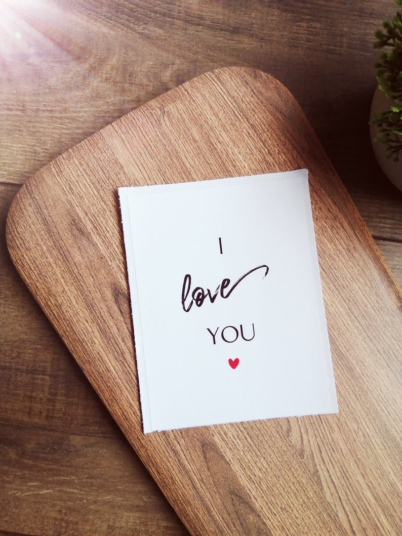 Pu&ograve; includere: Un biglietto bianco con testo nero che dice "Ti amo" e un piccolo cuore rosso.