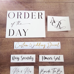 May include: A collection of white vinyl decals with black and gold lettering. The text includes ORDER OF THE DAY, initials AR, Custom Wedding Decal, Ring Security, Flower Girl, Mr & Mrs, and Bride To Be in various serif and script fonts.