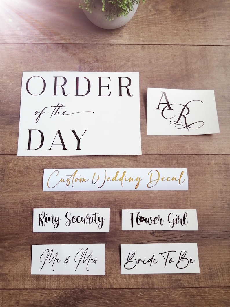 May include: A collection of white vinyl decals with black and gold lettering. The text includes ORDER OF THE DAY, initials AR, Custom Wedding Decal, Ring Security, Flower Girl, Mr & Mrs, and Bride To Be in various serif and script fonts.