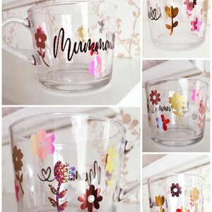 May include: A clear glass mug with a handle decorated with colorful flowers and glitter. The mug has the word "Mumma" written on it in black.