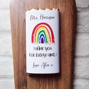 Op de afbeelding: Een chocoladereep met een regenboogontwerp en de tekst "Mr Harrison, THANK YOU FOR EVERYTHING! Love Alex x" op een houten serveerplank. De plank heeft een decoratieve rand.