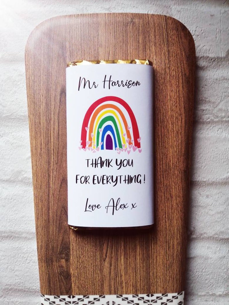 Op de afbeelding: Een chocoladereep met een regenboogontwerp en de tekst "Mr Harrison, THANK YOU FOR EVERYTHING! Love Alex x" op een houten serveerplank. De plank heeft een decoratieve rand.