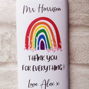Op de afbeelding: Een chocoladereep verpakt in wit papier met een regenboogontwerp en de tekst "Mr Harrison, Bedankt voor alles! Love Alex x." De regenboog heeft rode, oranje, gele, groene, blauwe en paarse bogen met roze hartjes.