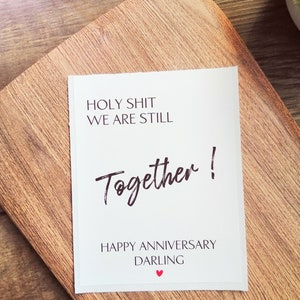 Pu&ograve; includere: Carta bianca con testo nero che dice "HOLY SHIT WE ARE STILL Together! HAPPY ANNIVERSARY DARLING" con un piccolo cuore rosso.