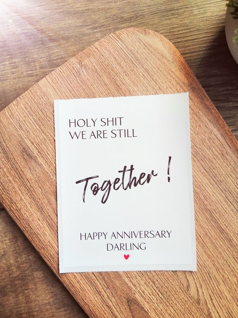 Pu&ograve; includere: Carta bianca con testo nero che dice "HOLY SHIT WE ARE STILL Together! HAPPY ANNIVERSARY DARLING" con un piccolo cuore rosso.