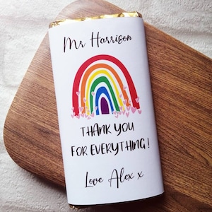 Op de afbeelding: Een witte chocoladereepverpakking met de tekst "Mr Harrison" boven een regenboogontwerp en de woorden "THANK YOU FOR EVERYTHING!" en "Love Alex x". De reep ligt op een houten oppervlak.