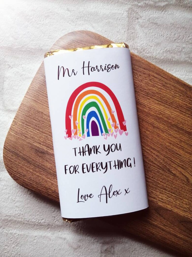 Op de afbeelding: Een witte chocoladereepverpakking met de tekst "Mr Harrison" boven een regenboogontwerp en de woorden "THANK YOU FOR EVERYTHING!" en "Love Alex x". De reep ligt op een houten oppervlak.