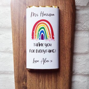 Op de afbeelding: Een chocoladereep met een regenboogontwerp en de tekst "Mr Harrison THANK YOU FOR EVERYTHING! Love Alex x" op een houten plank. De plank heeft een decoratieve rand.