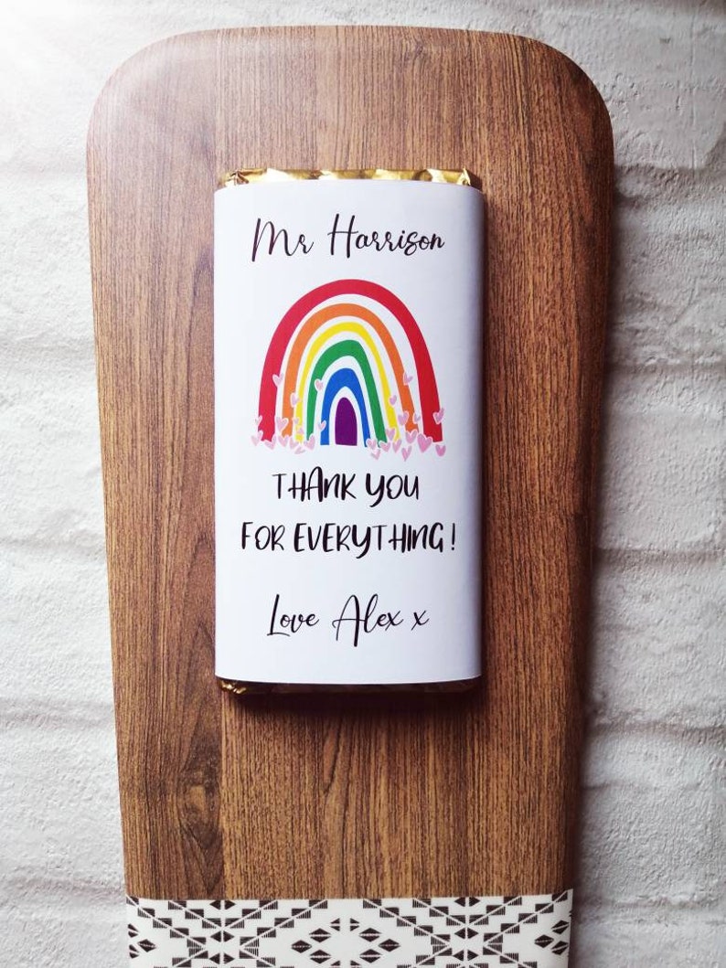 Op de afbeelding: Een chocoladereep met een regenboogontwerp en de tekst "Mr Harrison THANK YOU FOR EVERYTHING! Love Alex x" op een houten plank. De plank heeft een decoratieve rand.