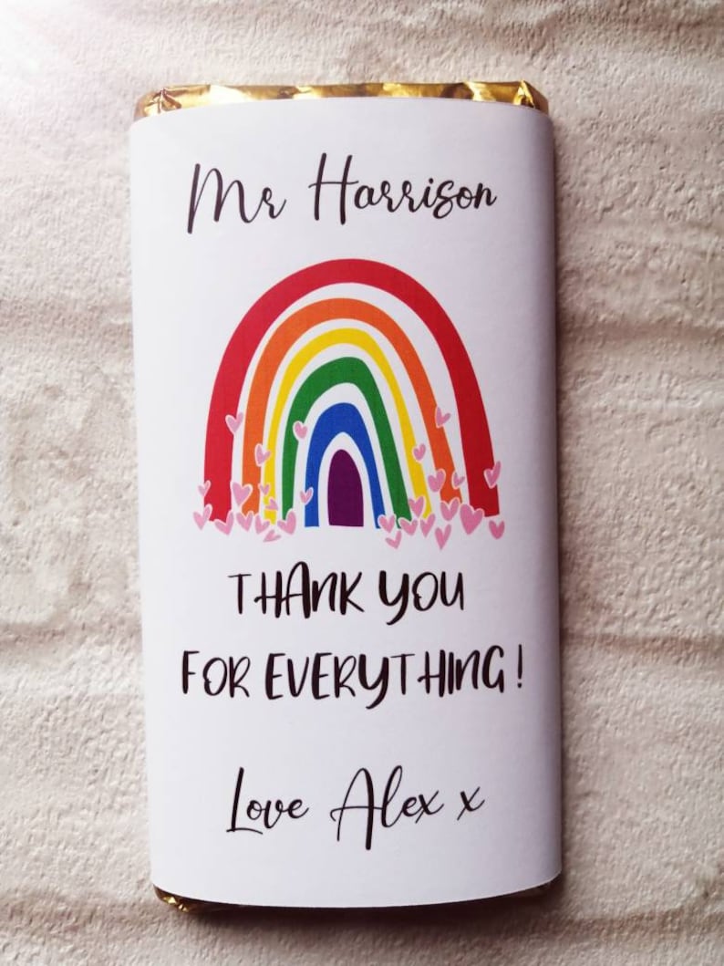 Op de afbeelding: Een chocoladereep verpakt in wit papier met een regenboogontwerp en de tekst "Mr Harrison, Bedankt voor alles! Love Alex x." De regenboog heeft rode, oranje, gele, groene, blauwe en paarse bogen met roze hartjes.