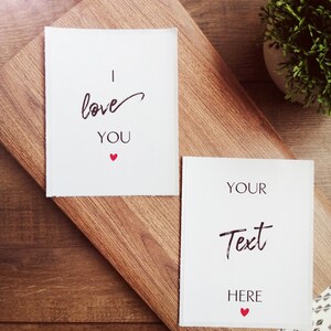 Pu&ograve; includere: Due carte bianche su una superficie di legno. La carta superiore ha il testo "Ti amo" con un piccolo cuore rosso. La carta inferiore ha il testo "Il tuo testo qui" con un piccolo cuore rosso.