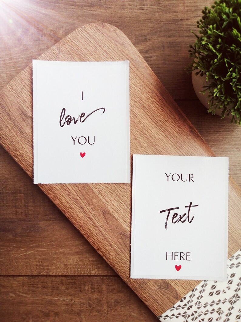 Pu&ograve; includere: Due carte bianche su una superficie di legno. La carta superiore ha il testo "Ti amo" con un piccolo cuore rosso. La carta inferiore ha il testo "Il tuo testo qui" con un piccolo cuore rosso.