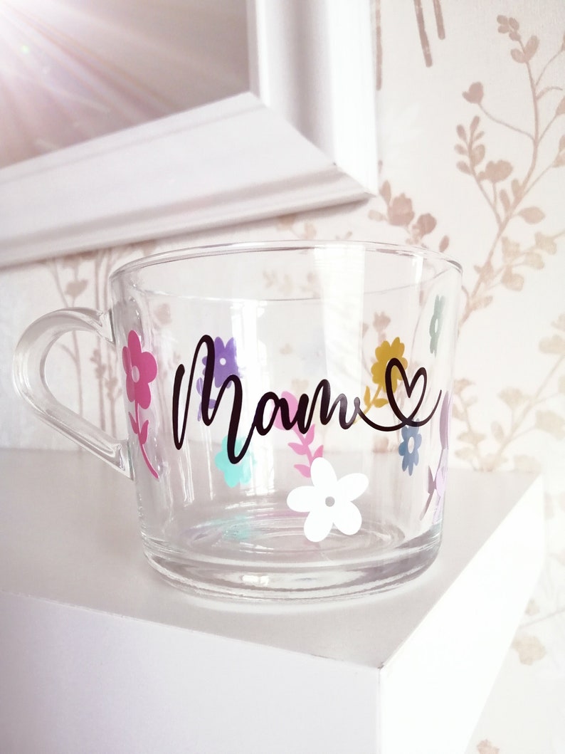 May include: A clear glass mug with a floral design and the word "Mom" in black lettering. The flowers are in shades of pink, purple, yellow, blue, and white.