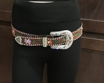 May include: Black leggings with a decorative belt. The belt has a brown strap with floral and rhinestone accents and a large silver buckle. The leggings are high-waisted and the belt is cinched around the waist.