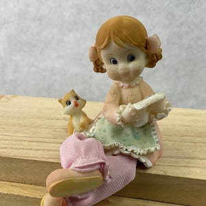 May include: A ceramic figurine of a girl with red hair and pink clothing, holding a white object. A small orange cat sits beside her. The figurine is on a wooden surface. The girl wears a green apron.