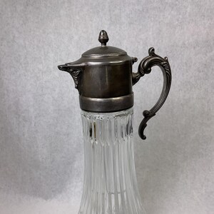 May include: A vintage clear glass pitcher with a silver-colored metal top, handle, and lid. The glass body has vertical ribbed detailing. The handle is ornate. The pitcher is likely for serving beverages.