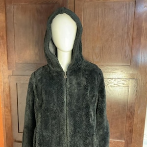 May include: A dark gray, hooded jacket with a zip-up front. The jacket has a soft, fuzzy texture and is displayed on a mannequin. The jacket's hood is up, and the mannequin is wearing black pants.