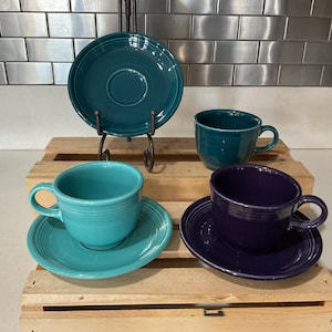 May include: A collection of ceramic dinnerware including one dark teal saucer on a metal stand, one dark teal cup, one light turquoise cup and saucer set, and one deep purple cup and saucer set. Each piece features a circular ridged design.