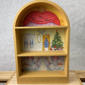 May include: A decorative, light yellow wooden shelf unit with three shelves. The top shelf features a red curtain with the text "The Nutcracker Ballet." The middle shelf has a Christmas tree and a chandelier. The bottom shelf has a candy cane design.