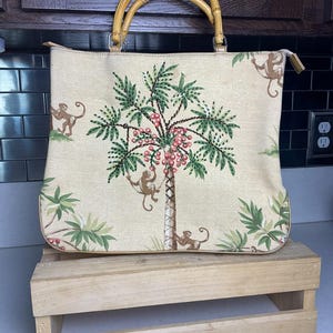 May include: A beige tote bag with a bamboo handle. The bag features a tropical design with palm trees, monkeys, and pink berries. The bag is sitting on a wooden crate.