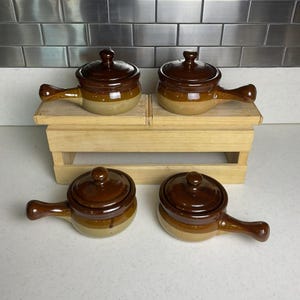 May include: Four small, brown and tan ceramic soup bowls with lids and handles. The bowls are arranged on a small wooden display stand. The lids have a small knob on top for easy handling. The bowls are ideal for serving individual portions of soup or stew.