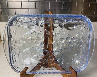 May include: A clear, rectangular glass baking dish with a blue rim and a fruit design. The dish is supported by a wooden stand. The fruit design includes grapes, apples, and other fruits.
