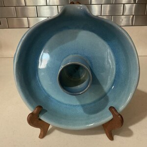 May include: A round ceramic baking dish with a central tube, featuring a smooth, glossy glaze in varying shades of light and medium blue. The dish has a slightly raised rim and a small pouring spout at the top edge, resting on a wooden stand.