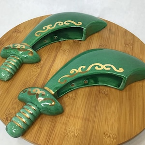 May include: Two green ceramic decorative pieces with gold accents, resembling stylized swords or handles, rest on a round wooden surface. The pieces have a curved blade-like shape with a decorative gold swirl pattern and a cylindrical handle.