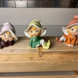 May include: Three ceramic figurines of elves with large hats and smiling faces. One elf wears a pink hat and brown outfit, another wears a green outfit and yellow hat, and the third wears an orange outfit and a peach hat.