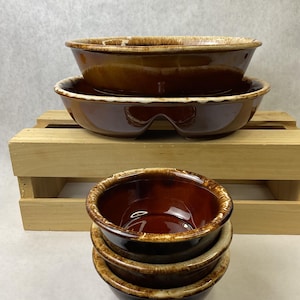 May include: A stack of four brown ceramic bowls with a glossy finish. The bowls have a two-tone design, with a lighter rim and a darker interior. Two larger bowls are stacked on a wooden crate.