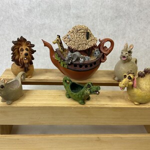 May include: A collection of animal figurines, including a lion, hippo, crocodile, camel, and rabbit, arranged on a wooden shelf. A brown ark-shaped teapot with animal figures on top is also present. The figurines are painted in various colors.