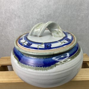 May include: A ceramic jar with a lid, featuring a white base with blue and green accents. The lid has a blue floral pattern and a curved handle. The jar is sitting on a wooden surface.