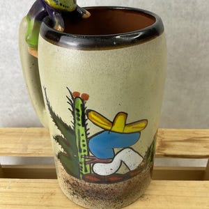 May include: A ceramic mug with a beige background and a brown rim. The mug features a painted cactus and a figure wearing a sombrero. The handle is a sculpted figure of a person in a sombrero. The mug is sitting on a wooden surface.