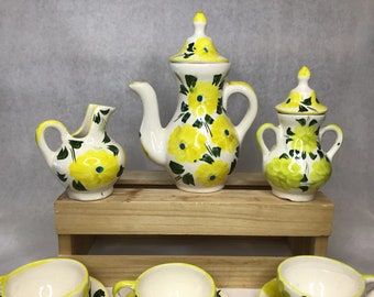 May include: A vintage ceramic tea set with a floral design. The set includes a teapot, creamer, sugar bowl, and cups, all decorated with yellow flowers, green leaves, and blue accents. The pieces are arranged on a wooden shelf.