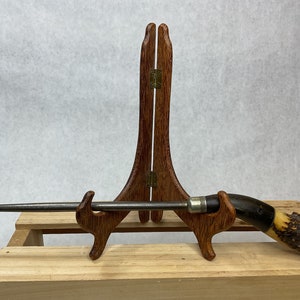 May include: A steel knife sharpener with a dark wooden handle and a silver metal band. The handle is made of dark brown wood and a lighter-colored material. The sharpener is displayed on a wooden stand.