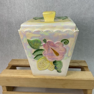May include: A square, iridescent ceramic jar with a lid, featuring a floral design with pink flowers, green leaves, and yellow lemons. The lid has a yellow handle. The jar is set on a small wooden crate.