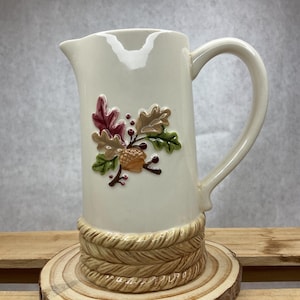 May include: Cream-colored ceramic pitcher with a decorative handle and spout. The pitcher features a raised relief design of autumn leaves and an acorn. The base has a woven rope design. The pitcher is displayed on a wooden slice.
