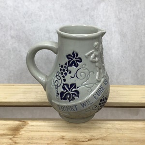 May include: Gray ceramic pitcher with a handle and spout. The pitcher is decorated with blue grape and leaf designs, and a relief of a figure. The words "TRINKT WIE EURE VATER" are inscribed around the base.