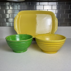 May include: A set of three ceramic kitchen items: a large yellow rectangular serving dish and two bowls, one green and one yellow. The bowls have a ribbed design. The items are arranged on a white surface.